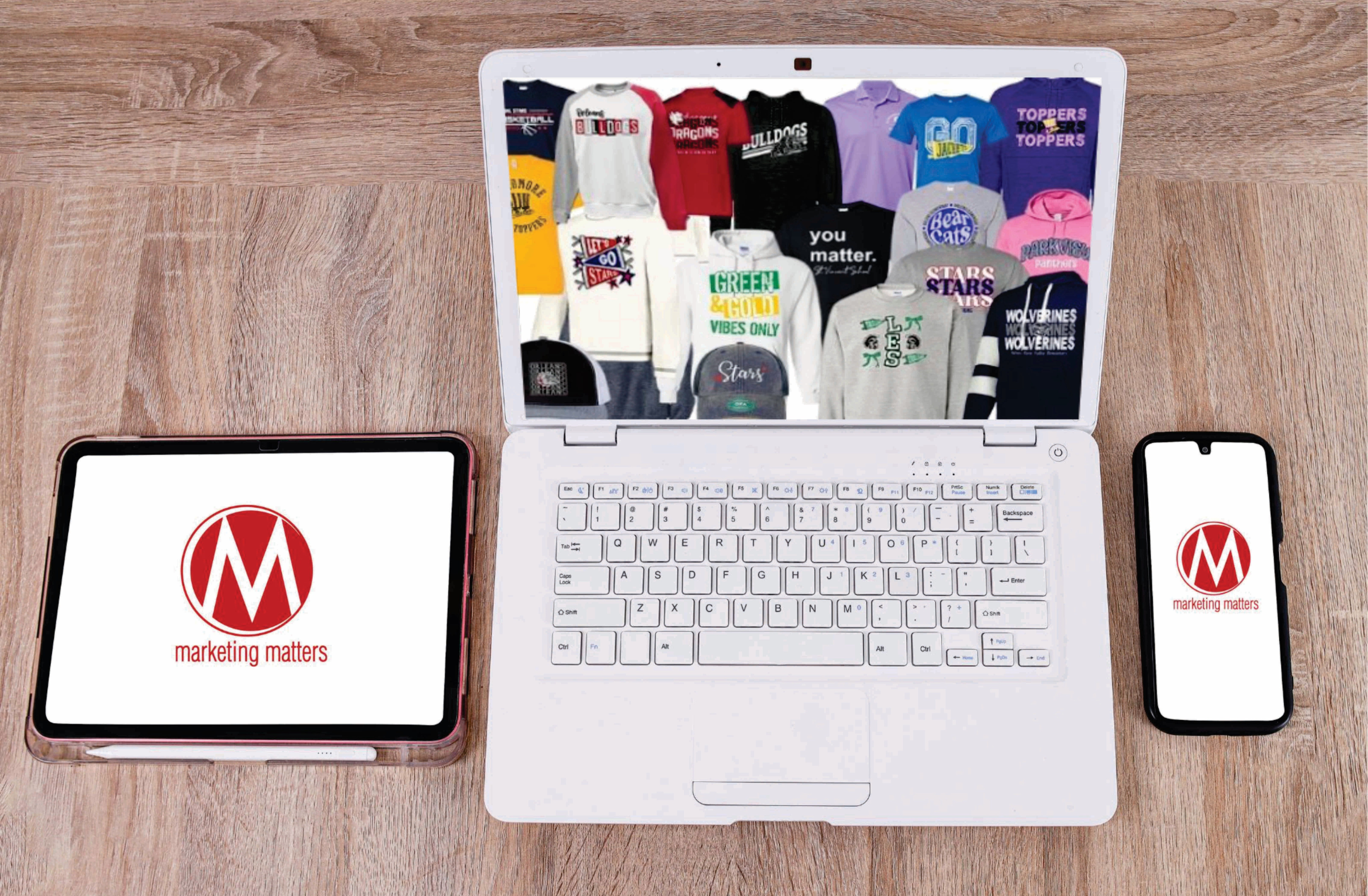 Laptop displaying various printed t-shirts, tablet, and phone with 'marketing matters' logo on a wooden surface.